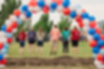 Six people in colorful shirts stand in a field under a red, white, and blue balloon arch. Trees and a crowd are visible in the background.