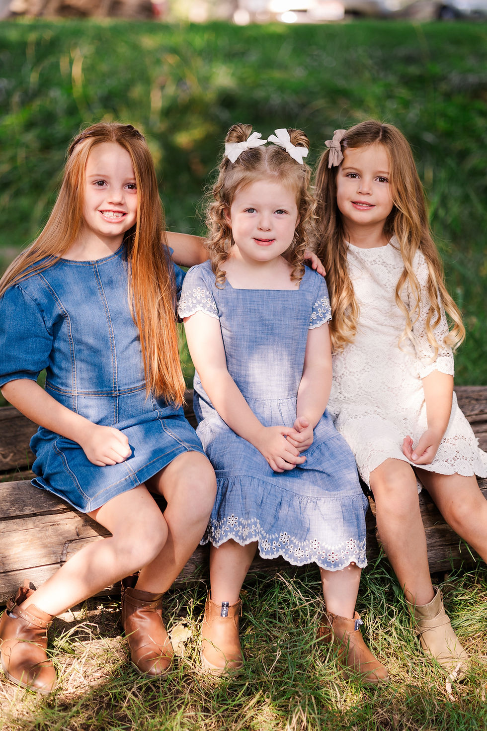 Three young girls sit on a log outdoors, wearing denim and lace dresses with boots. They smile warmly, surrounded by green grass.