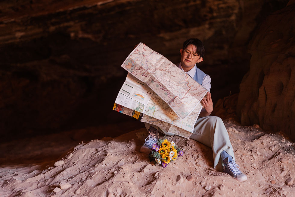 Person in a cave studying a large map, wearing light-colored clothing. Nearby, a bouquet of colorful flowers lies on the sandy ground.