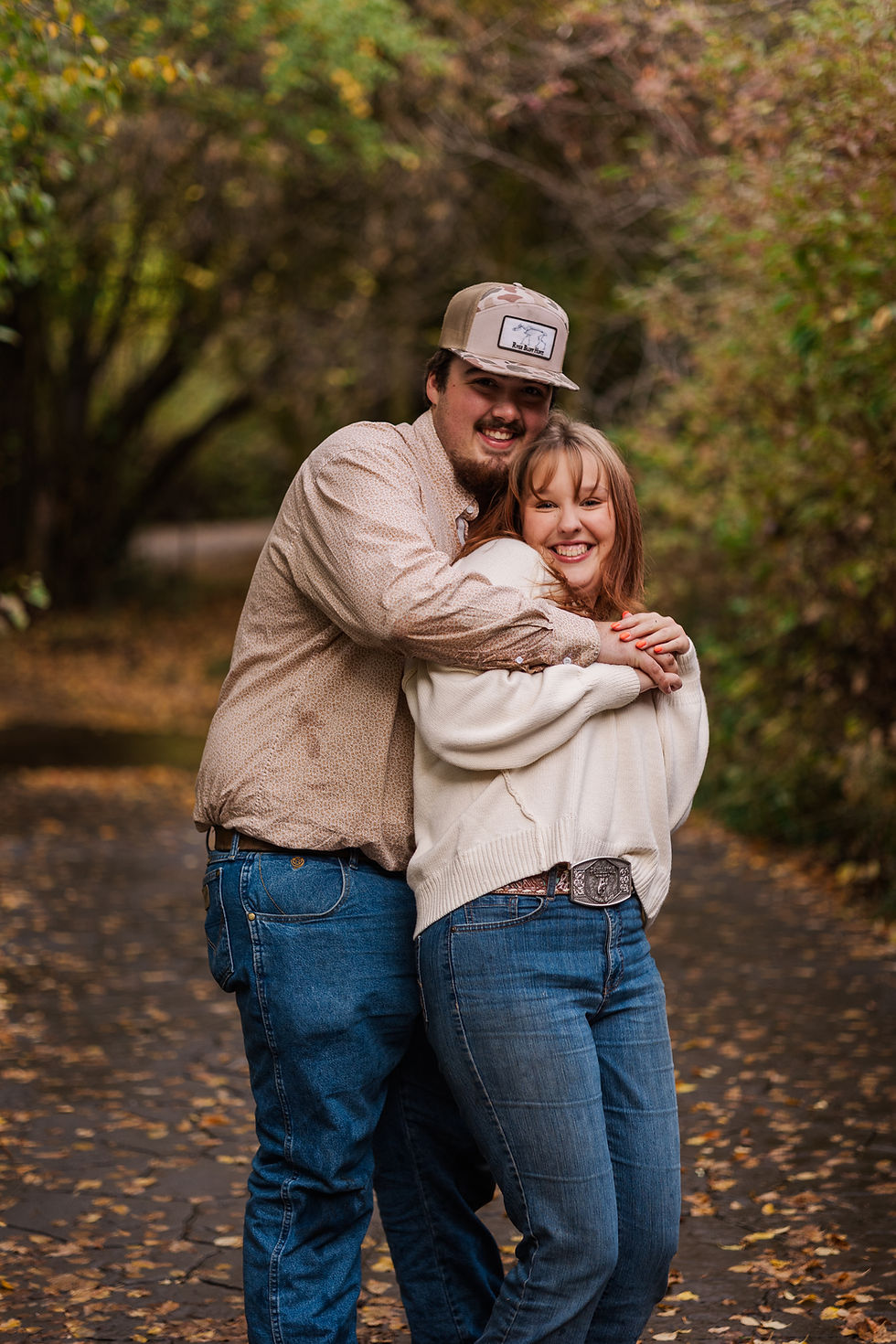 A couple embraces happily on a leaf-covered path in a park. They wear casual clothes, with autumn leaves and green trees in the background.