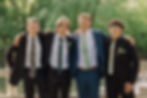 Four men in suits, smiling with arms around each other, pose outdoors on a wooden bridge. Greenery in the background suggests a sunny day.