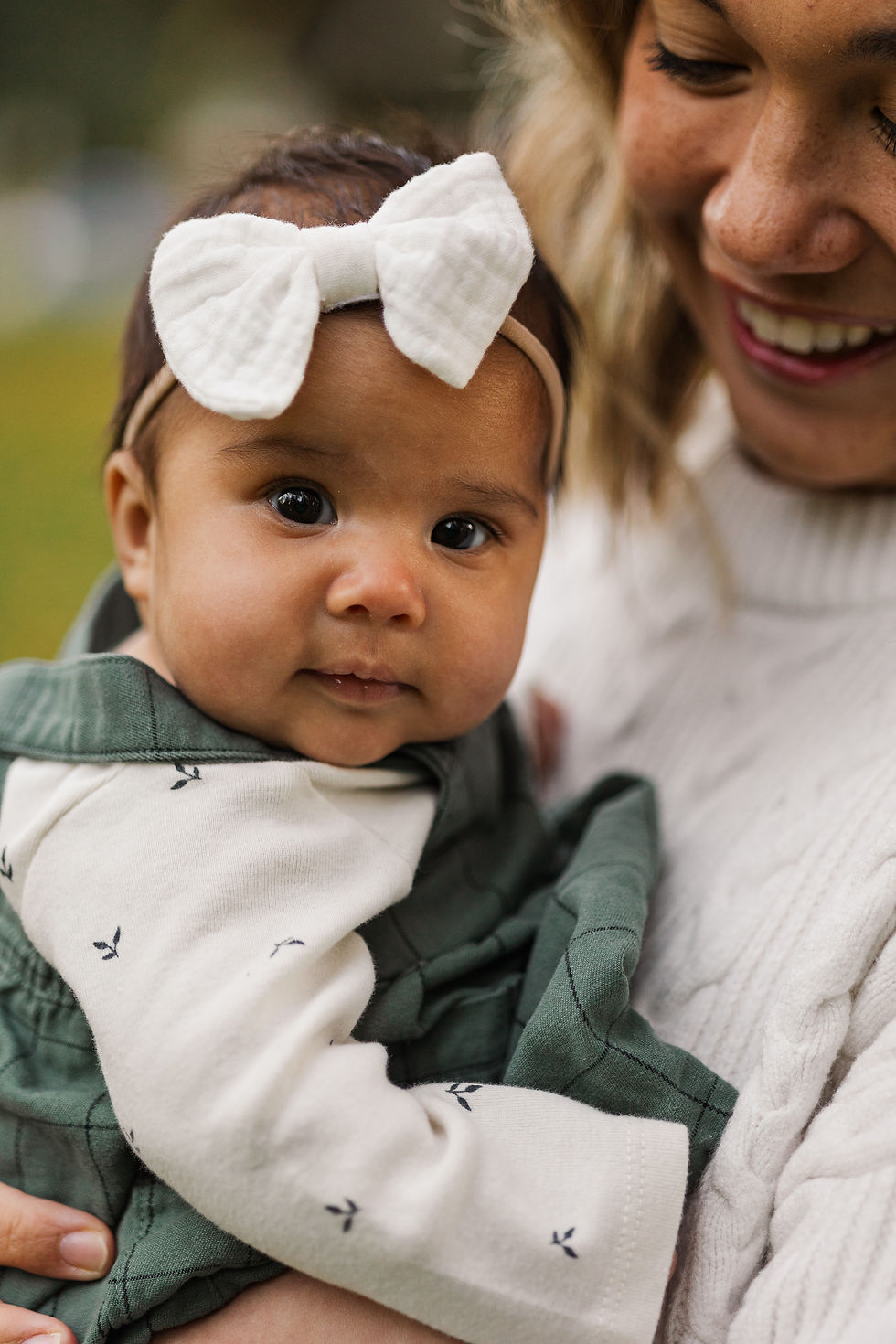 Smiling woman holds a baby with a white bow. The baby wears a green outfit with a pattern. Soft, warm setting, close-up shot.