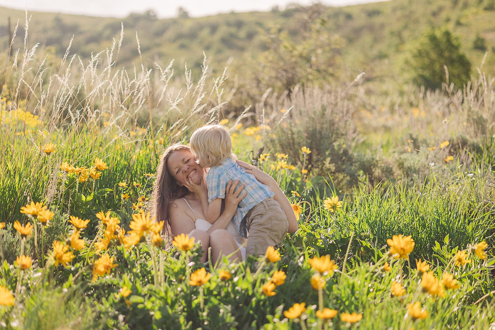 Woman and child embrace joyfully in a meadow with yellow flowers. Sunny day, green hills in the background. Both appear happy and playful.