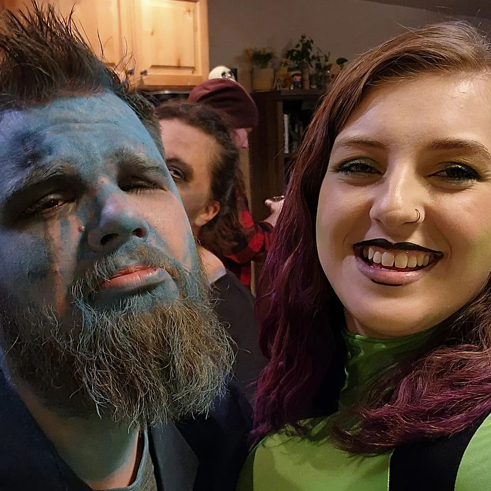 Man with blue face paint and woman with purple hair smiling in a cozy room. Wood cabinets in background. Playful atmosphere.