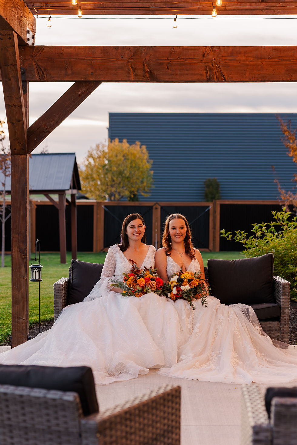 Two women in white gowns sit on a sofa holding colorful bouquets. They are outdoors under a wooden pergola with string lights, looking happy.