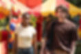 Two people stand in front of a colorful carnival tent with balloons. One wears a yellow shirt, the other a brown shirt, both appear focused.