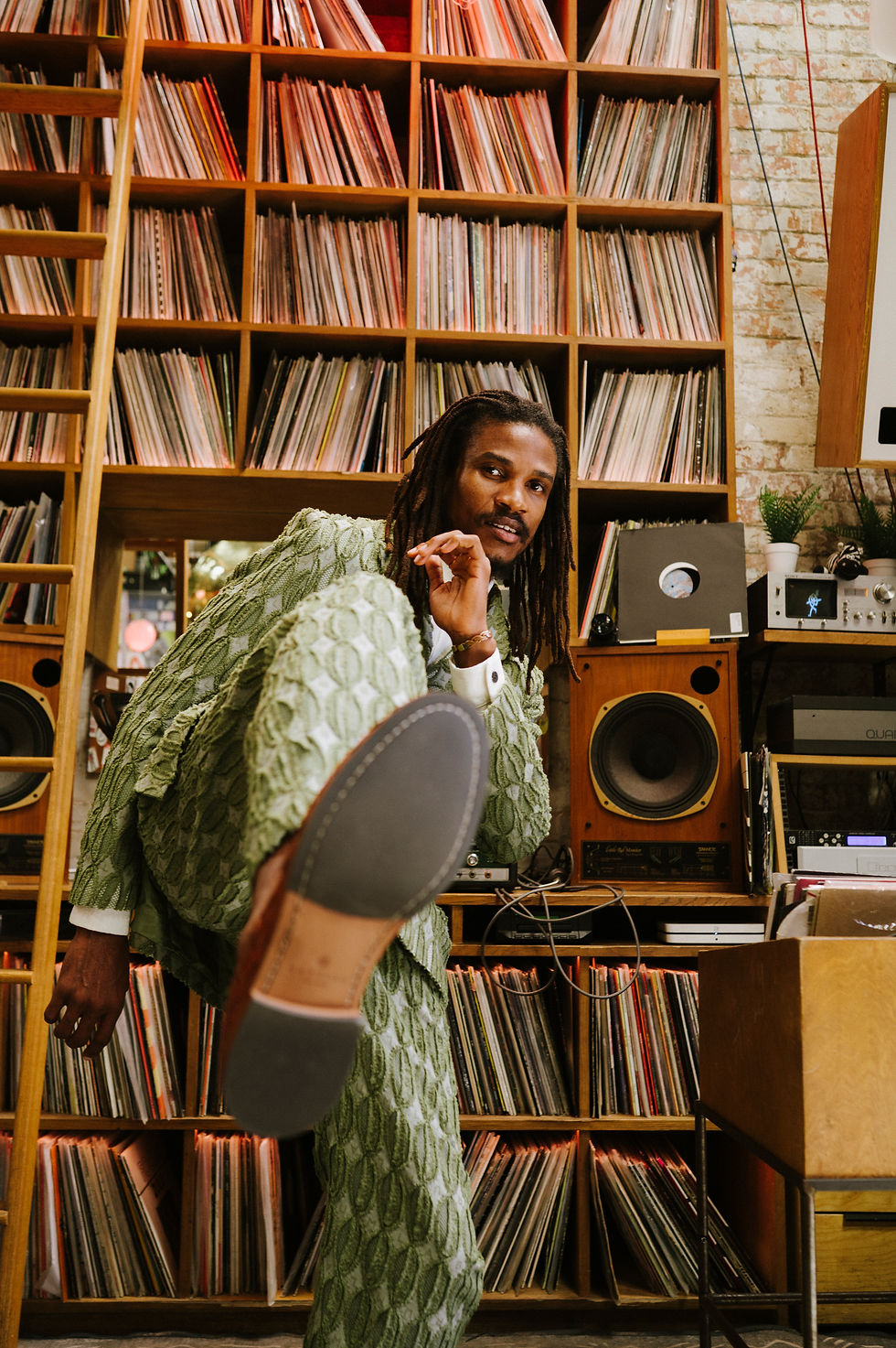 Man in a green suit kicks his foot towards the camera, in the background there is a wall of vinyls and a large speaker