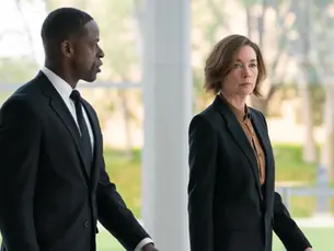 Two people in suits walk indoors, appearing serious. Large windows reveal a blurred green garden outside. Bright, formal setting.