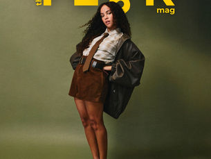 Actor Lou Llobell poses in front of a green backdrop. She wears a brown suede style skirt, an oversized bomber jacket and a shirt with a brown tie. 
