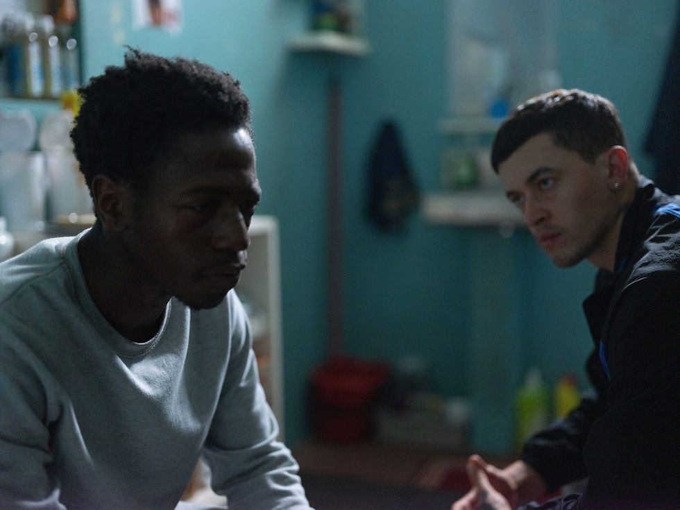 Two men in sit in close quarters, the one in the forefront is wearing a grey long sleeved jumper whilst the one in the background is wearing a dark jacket.