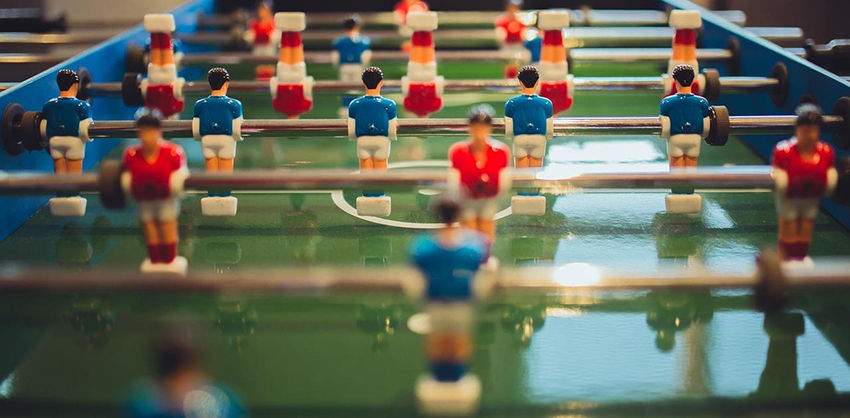 Blue and red foosball players on a green field. Metal rods connect figures. Bright colors, no text, indoor setting, playful mood.