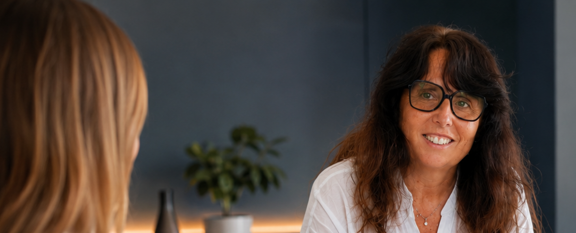 Deux femmes discutent à un bureau moderne; l’une sourit et écrit dans un carnet, avec des papiers et une plante en arrière-plan.