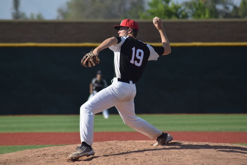A right handed pitcher in a black jersey with white stripes raises his arm behind his head as he prepares to throw a pitch