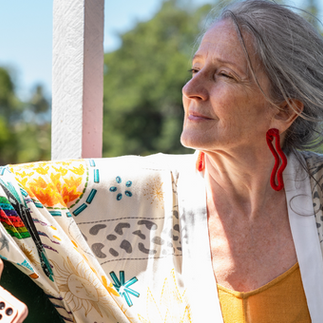Eine Frau,, graue Haare, Weiße Jacke mit bunten Mustern und gelbes Top sitzt entspant und schaut in die Ferne.