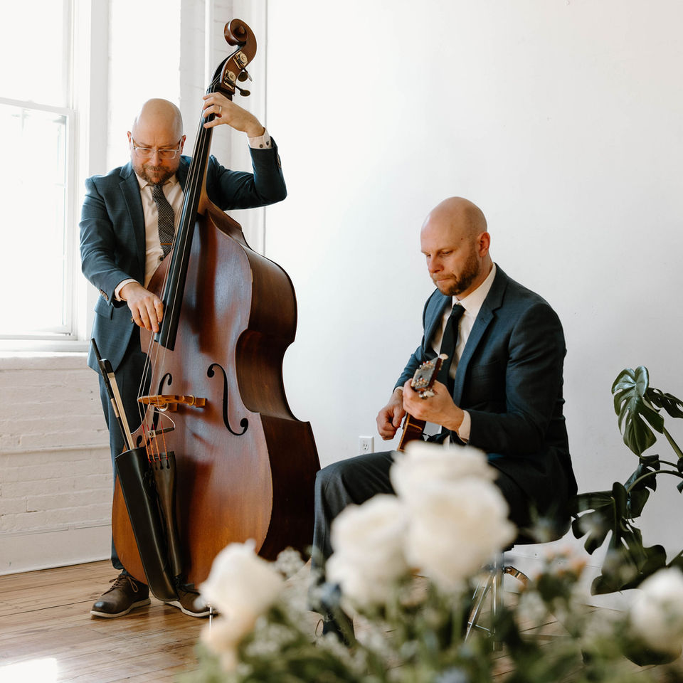 flower arrangement blurred out in the foreground, with jazz duo playing music in focus in the background. Bass player and guitarist are wearing matching suits.