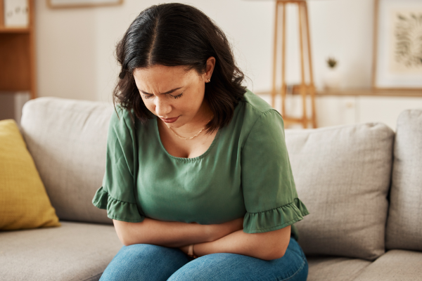 Woman in a green shirt sits on a beige couch, clutching her stomach and looking in pain.