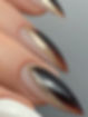 Close-up of fingers with long, pointed nails featuring a black, brown, and gold glitter ombre design. The mood is elegant and stylish.