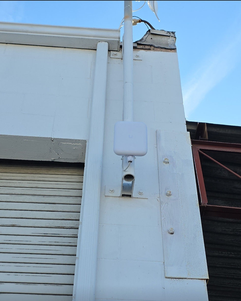 White wifi access point mounted on a white industrial building wall. Blue sky in the background. Metal roof support visible. Calm setting.