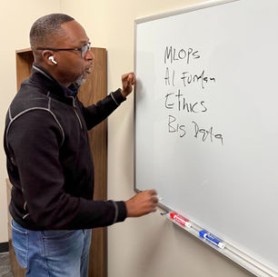 Dr. Ogunlana standing beside a whiteboard explaining MLOps, AI foundations, ethics, and big data during a responsible AI and cybersecurity instruction session.