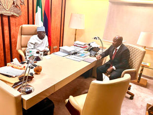 President Bola Ahmed Tinubu, dressed in a white traditional agbada and cap, sits behind his desk in his office, speaking with Oluwatosin Ajayi, Director General of the State Security Service, who sits across from him in a dark suit and red patterned tie. The Nigerian flag and the national coat of arms are visible behind the President, and documents are stacked on the desk between them.