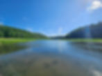 Calm "lake" with lush green hills under a bright blue sky with scattered clouds. Sunlight creates a serene and peaceful atmosphere.