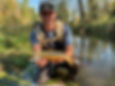 Man kneels by a stream holding a Brook Trout. He wears a backpack and sunglasses, surrounded by green foliage, under a clear blue sky.