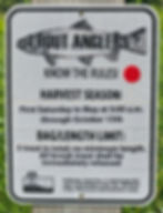 Sign for trout anglers with harvest season dates and rules on catch limits. Features a fish illustration. Background: greenery.