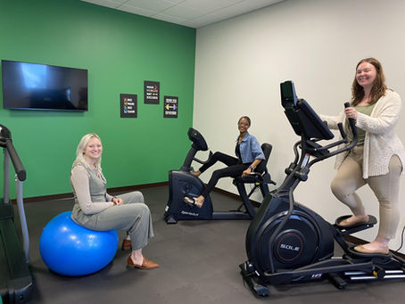 Three people in a gym: one on a blue exercise ball, another on a stationary bike, and one on an elliptical. Green wall with motivational signs.