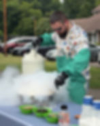 Man in apron and gloves makes a smoky dry ice treat outdoors, with colorful toppings and cups on a table by parked cars