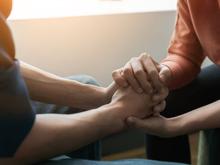 Two people are sitting closely, holding hands in a comforting manner. One wears an orange shirt, with soft sunlight in the background.