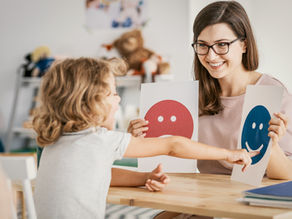 Child pointing at a blue happy face card held by a smiling woman in glasses. They sit at a table in a playroom with toys in the background.