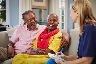 An elderly couple sits smiling on a sofa, engaged in conversation with a woman taking notes. Bright room, colorful outfits create a warm mood.