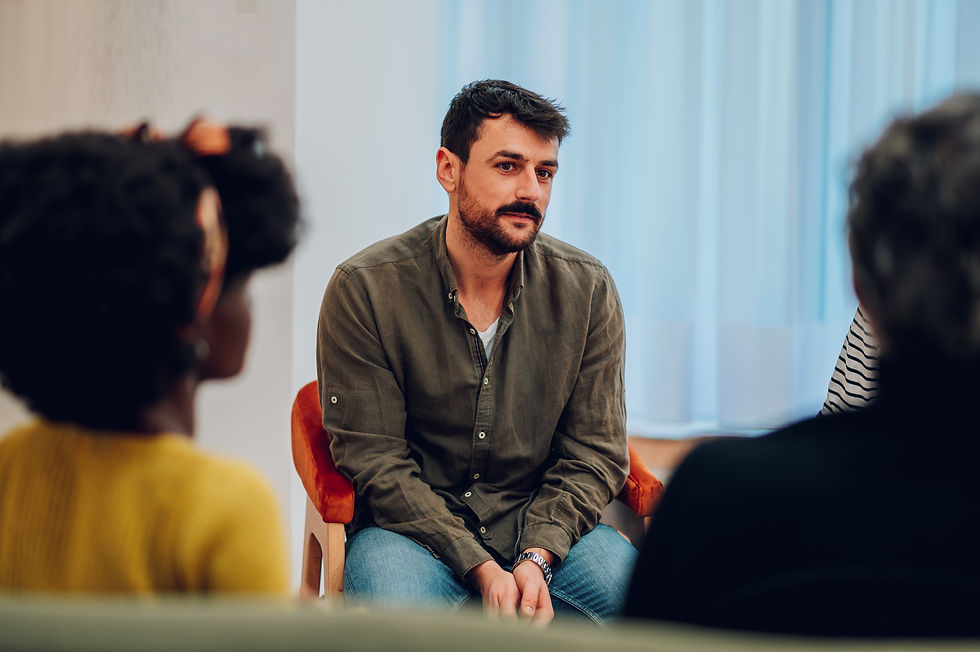 Man in a green shirt sits in a circle with others, appearing thoughtful. Blue curtain background, warm lighting, casual setting.