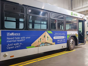 City bus in a garage with an ad for Alliance Healthcare Services, promoting mental health help and 24/7 walk-in services.