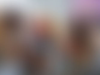 A teacher in a red polka dot shirt sits with five attentive children in a classroom, with colorful papers on a bulletin board behind them.