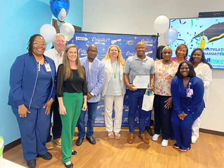 Group of people smile in a room with blue walls, balloons, and a "Congrats" banner. One holds a certificate. Celebratory mood.