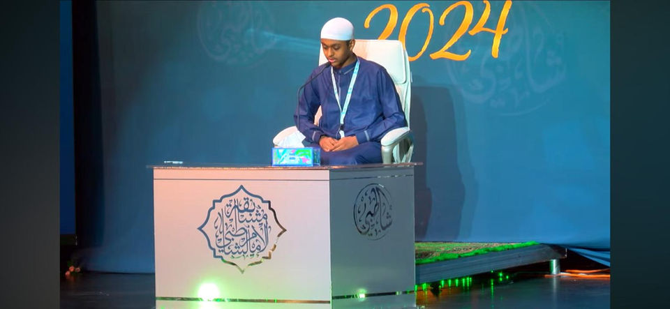 Man in blue shirt standing at podium with Arabic design, 2024 Dar Al Quran