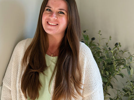 Smiling Hannah Morgan, Resident in Counseling, with long brown hair in a cream cardigan stands indoors beside a leafy plant against a beige wall.