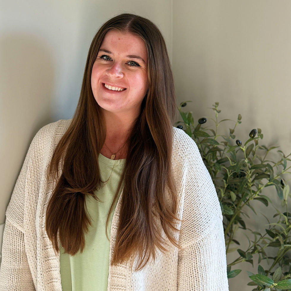 Smiling Hannah Morgan, Resident in Counseling, with long brown hair in a cream cardigan stands indoors beside a leafy plant against a beige wall.