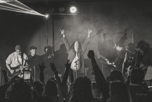 Black-and-white concert: a singer with arms raised, guitarists beside, and a crowd with hands up in stage haze