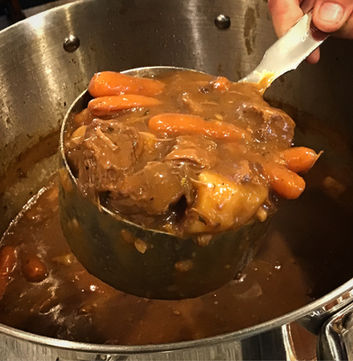 A ladle of beef stew held over the pot showing chunks of beef, carrots and potato in a rich brown gravy.
