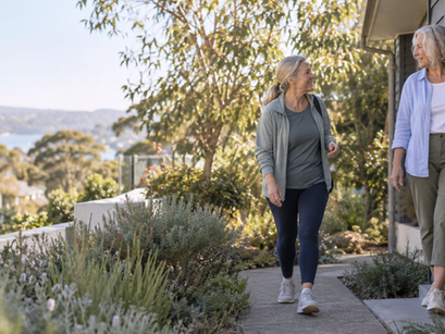 Two older women smile and chat while walking by a house garden with trees and a distant water view.
