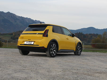 Renault 5 Elektro auf einem Parkplatz. Im sind Wald und Berge zu sehen.