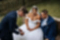 A bride in a white dress with a tiara and veil, her new husband in a blue suit and tie. They smile as they watch a card trick performed by Jamie. Jamie wears a blue suit and white shirt.