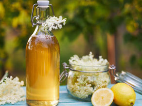 Sklenená fľaša s fermentovanou bazovou limonádou, zaváraninový pohár s bazovými kvetmi a rozkrojený citrón na blog o letnom osviežení.