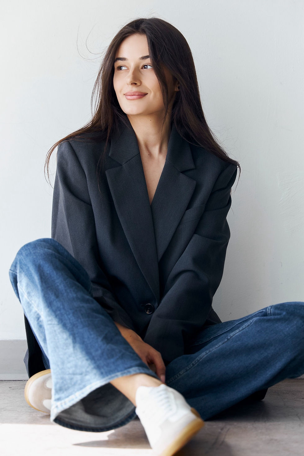Vladyslava Kostovska seated casually in a gray blazer and jeans, with a confident yet relaxed demeanor in a bright studio set