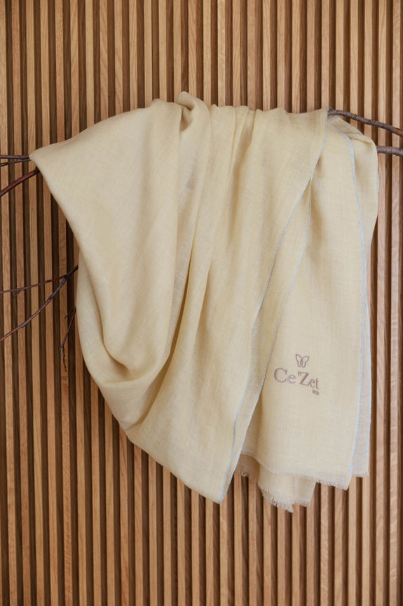 Beige Schal mit Ce'Zet-Logo, hängend vor Holz Hintergrund. Ce'Zet.