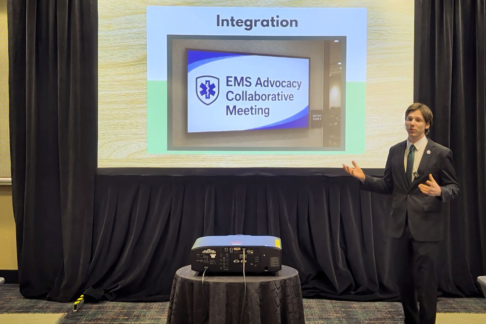 Man in a suit presents at the American Ambulance Association Annual Conference. On the projector screen is an image of a TV screen with the text "EMS Advocacy Collaborative Meeting" on it. Projector and black curtains set the conference room scene.