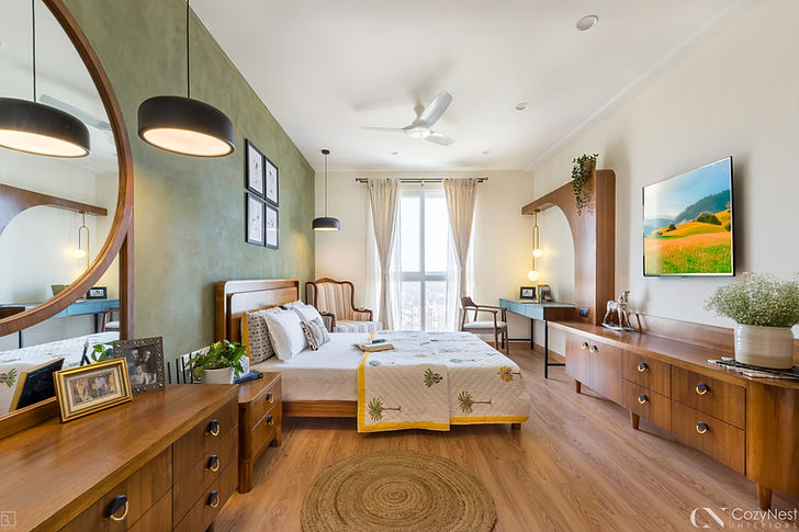 Modern bedroom interior design with elegant wooden furniture, a green accent wall, and warm natural lighting. The space showcases a stylish bed with patterned bedding, a dresser with a large round mirror, and a wall-mounted TV unit for functional luxury. Pendant lights, framed artwork, and indoor plants enhance the décor, while sheer curtains over the large window create an airy, sunlit vibe. This luxury bedroom interior design blends comfort and aesthetics, offering inspiration for modern bedroom décor ideas, cozy apartment interiors, and contemporary urban home styling. Perfect for homeowners searching for creative bedroom design inspiration with a balance of functionality and elegance.