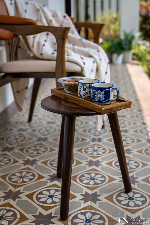 Close-up of a small, dark wooden three-legged side table on an outdoor patio featuring grey and blue geometric patterned cement tiles. The table holds a light wooden tray with two blue floral patterned mugs and a clear bowl, next to a chair with a person sitting under a blanket.